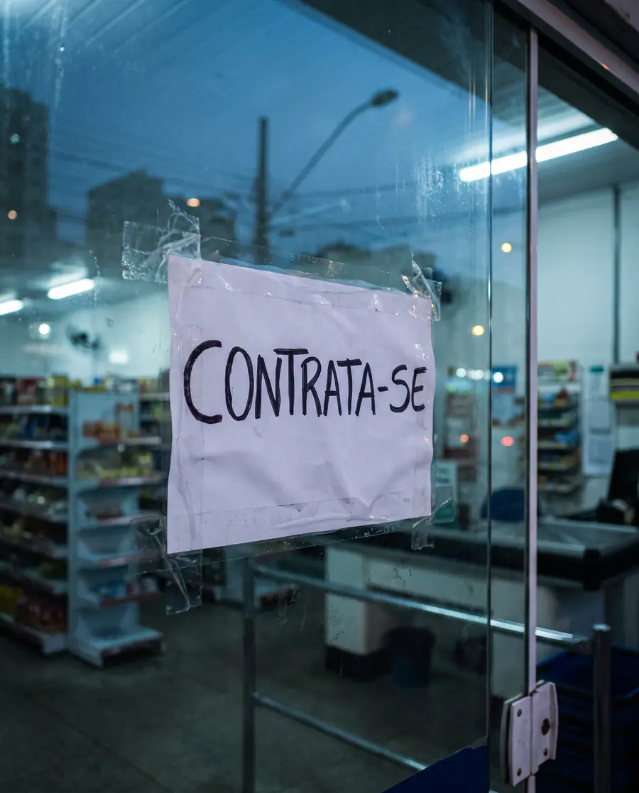 Folha com a palavra Contrata-se colada na porta de vidro de um supermercado, ao entardecer